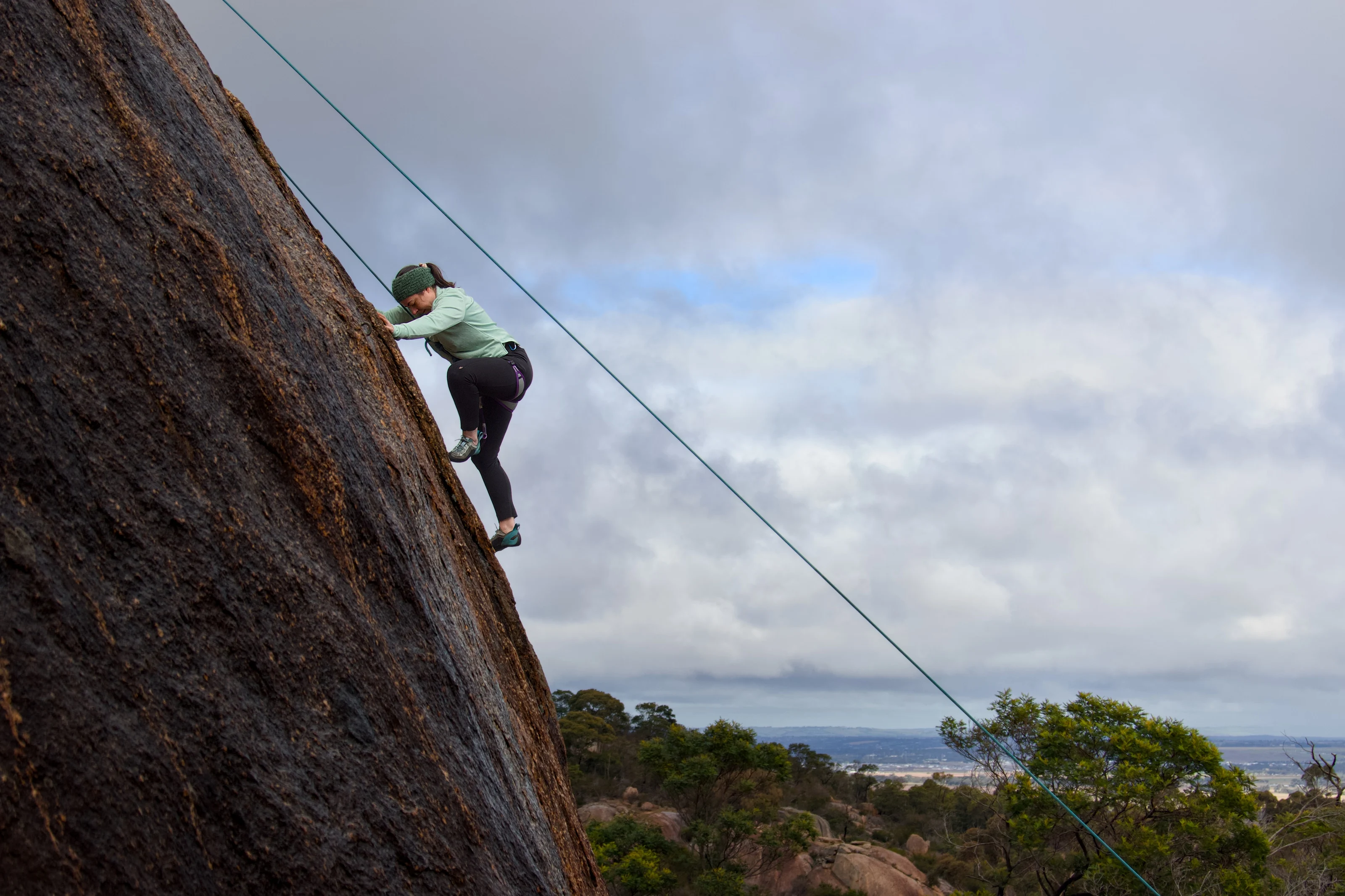 A climber in a pale green top climbs a dark rock face beneath a wide cloudy sky.