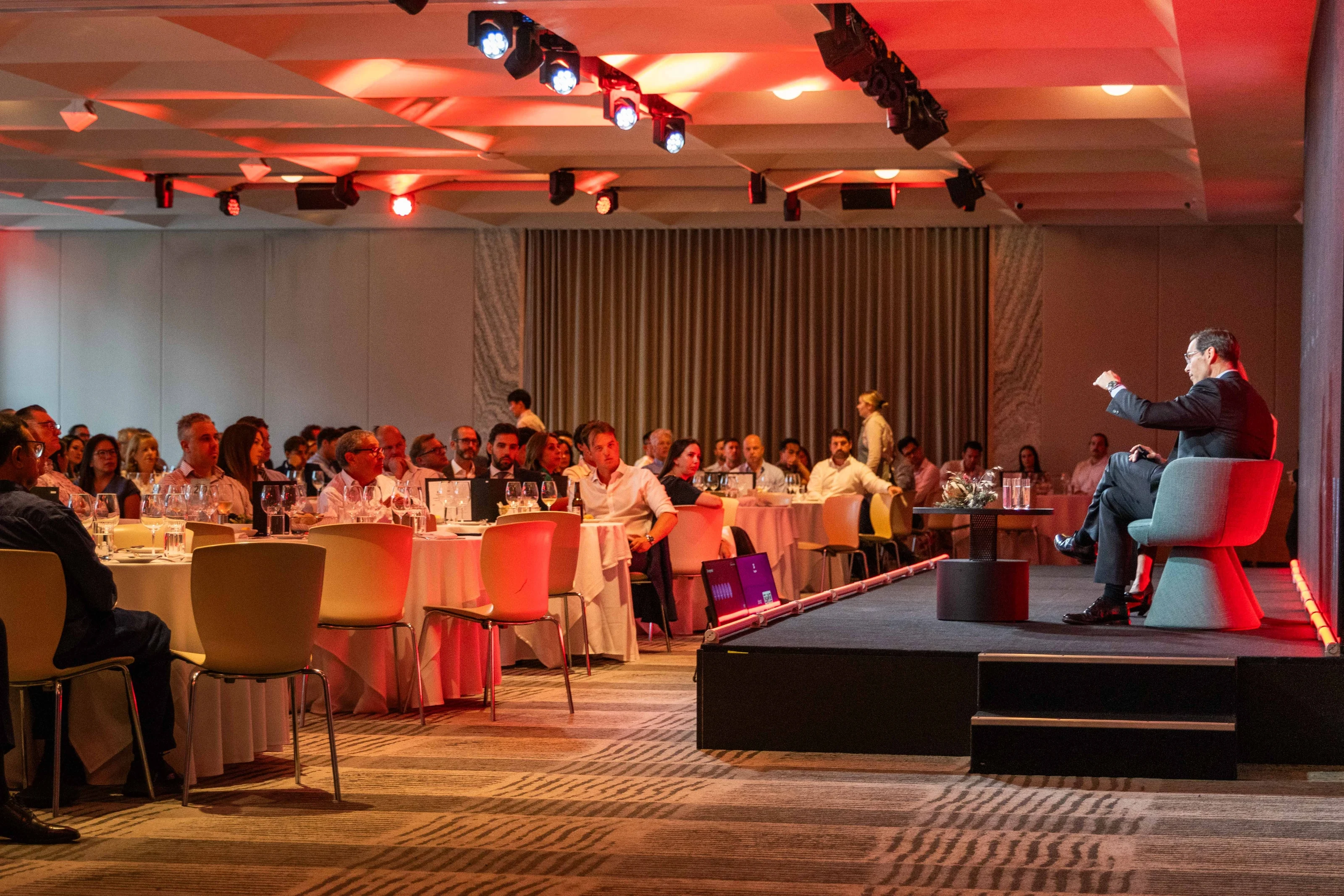 A seated speaker addresses guests around tables in a warmly lit event room.