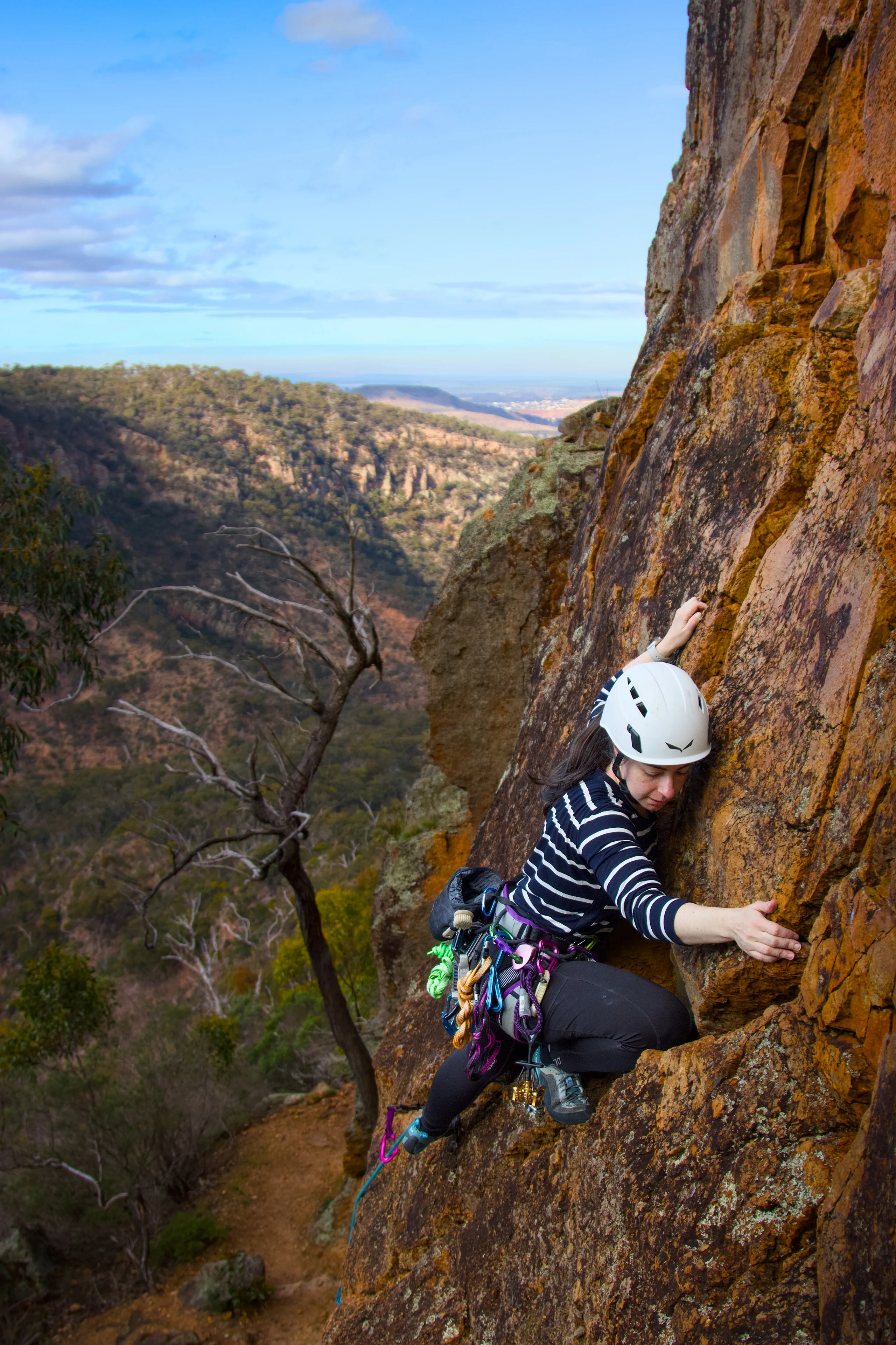 A climber in a white helmet and striped top holds orange rock above a wooded valley.