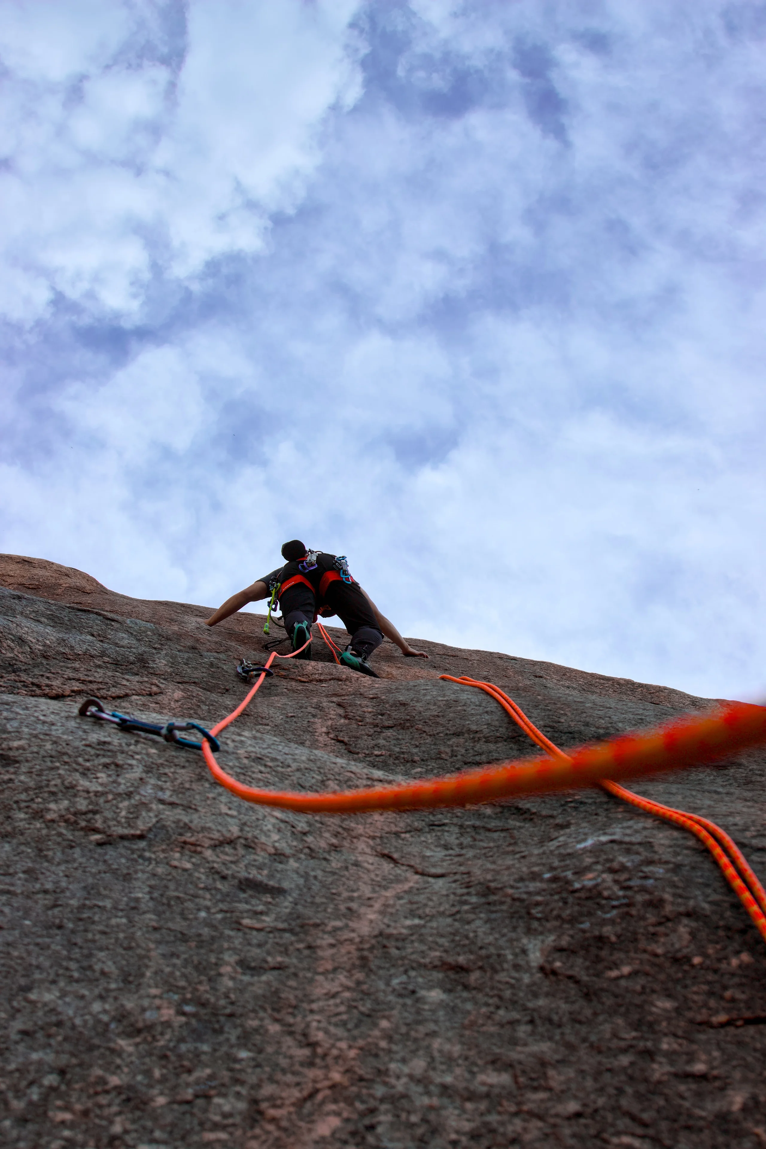 Orange ropes lead up a steep rock face to a climber beneath a cloudy sky.
