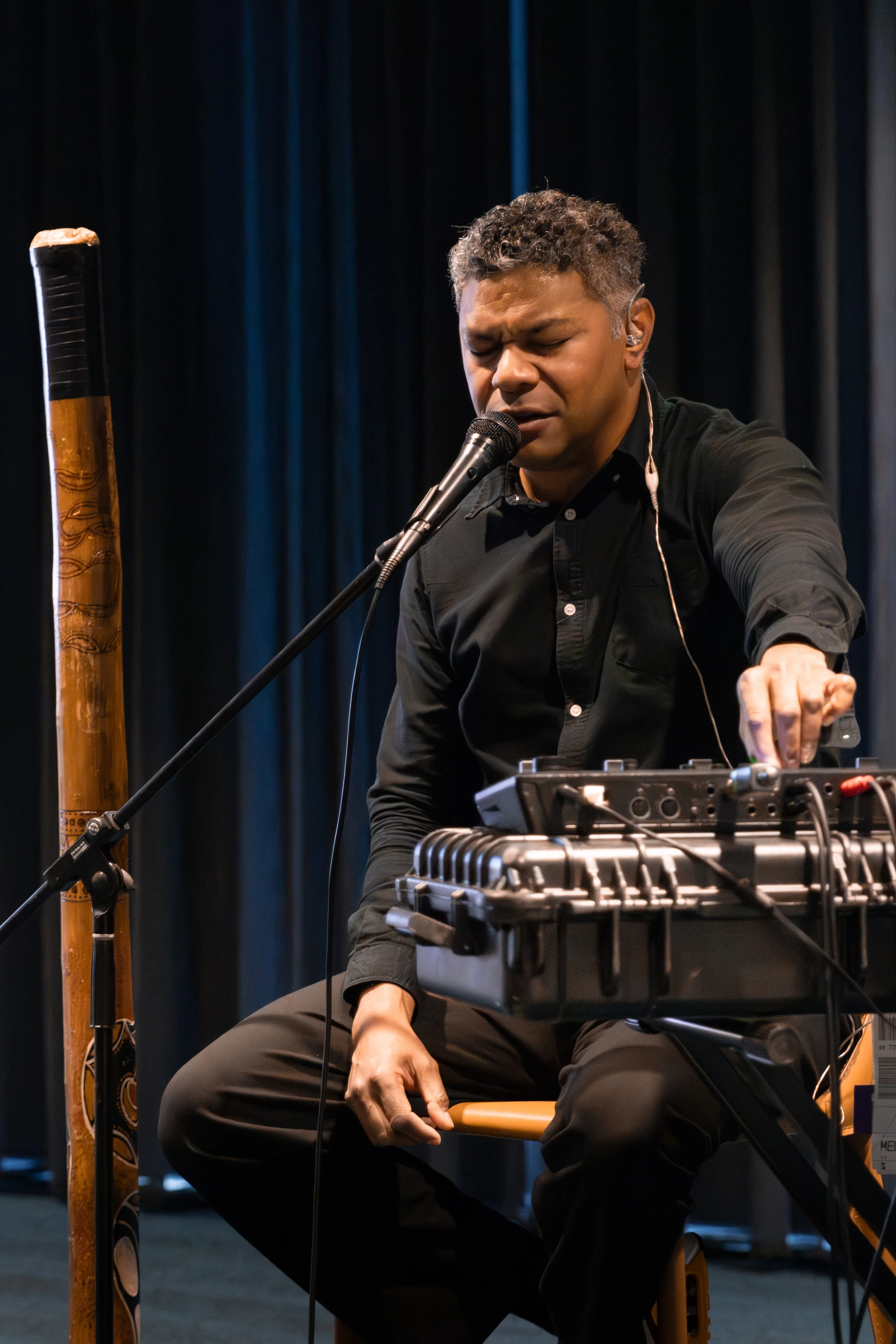 A seated musician sings into a microphone while adjusting audio equipment.