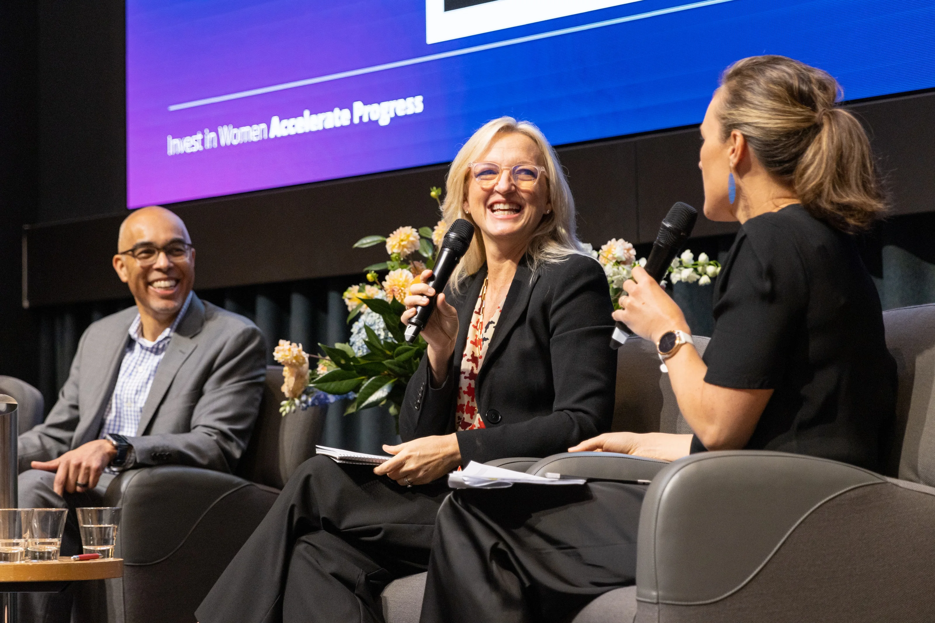 Three panellists smile and exchange remarks while holding microphones.