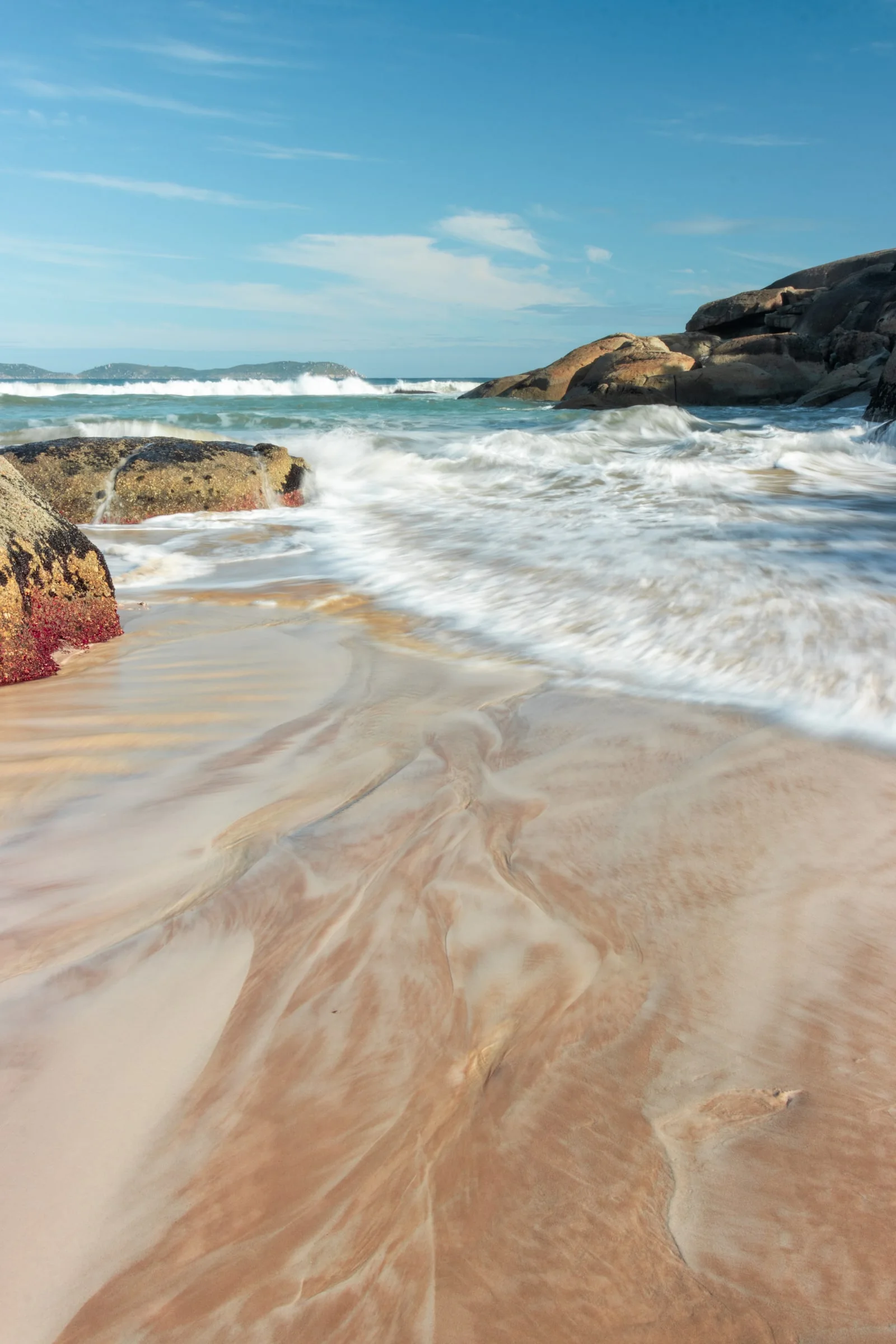 Long-exposure waves wash around granite rocks beneath a blue sky.