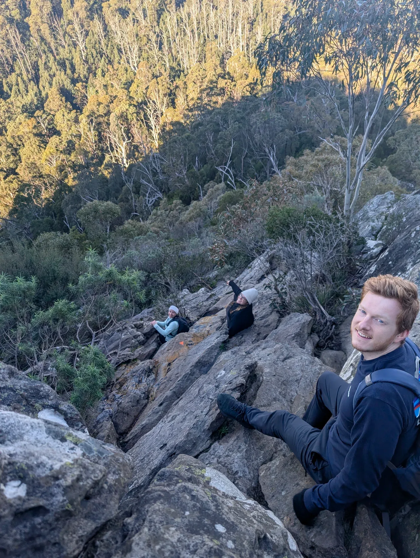 Three hikers pausing on a rocky slope above a wooded valley.