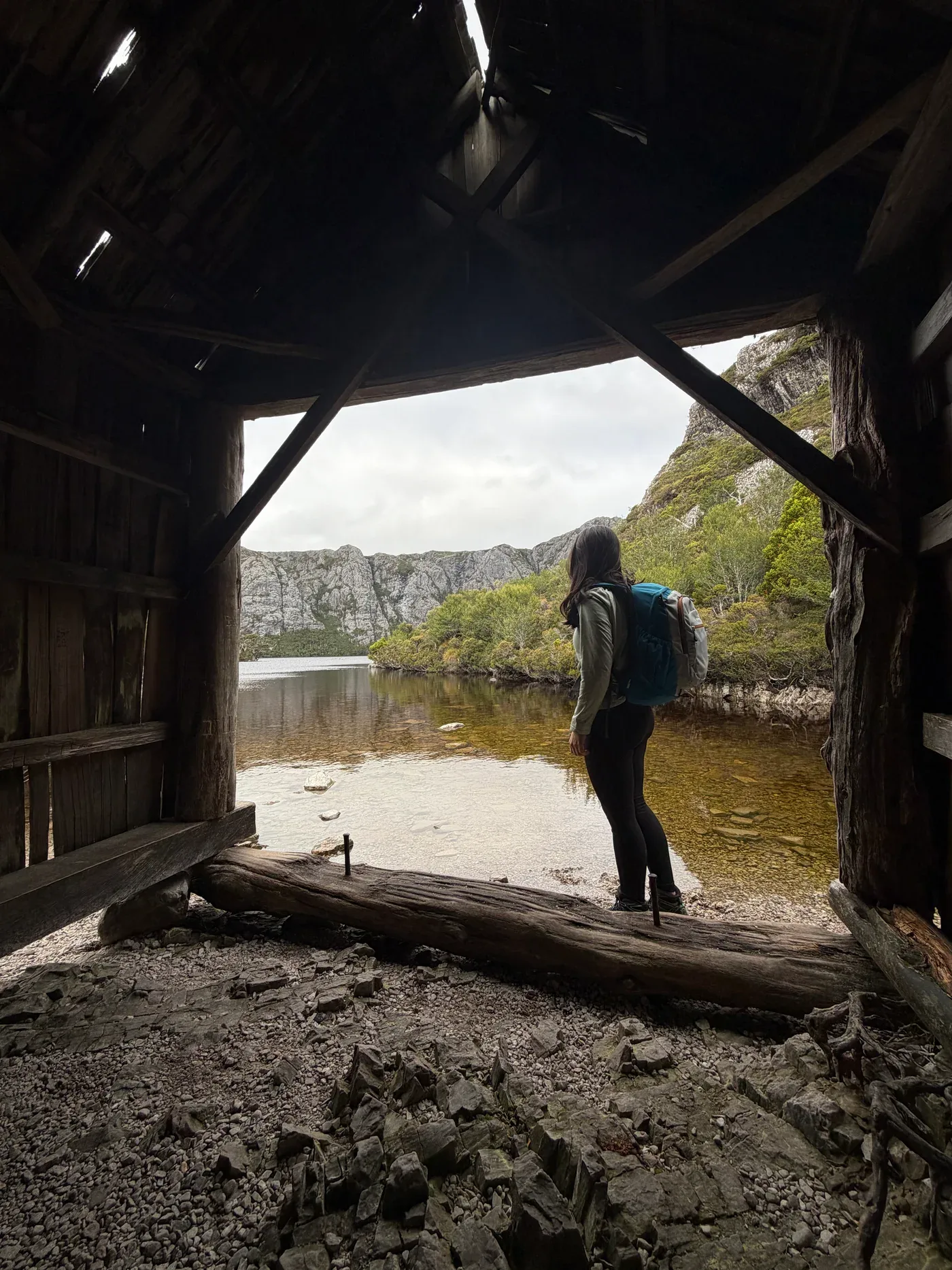 A hiker looking across a mountain lake from a timber shelter.