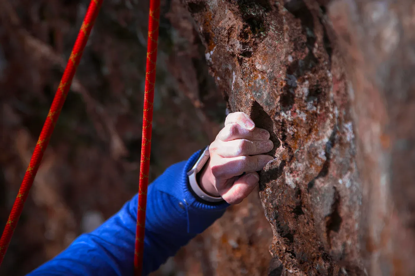A chalk-covered hand gripping textured rock beside a red climbing rope.